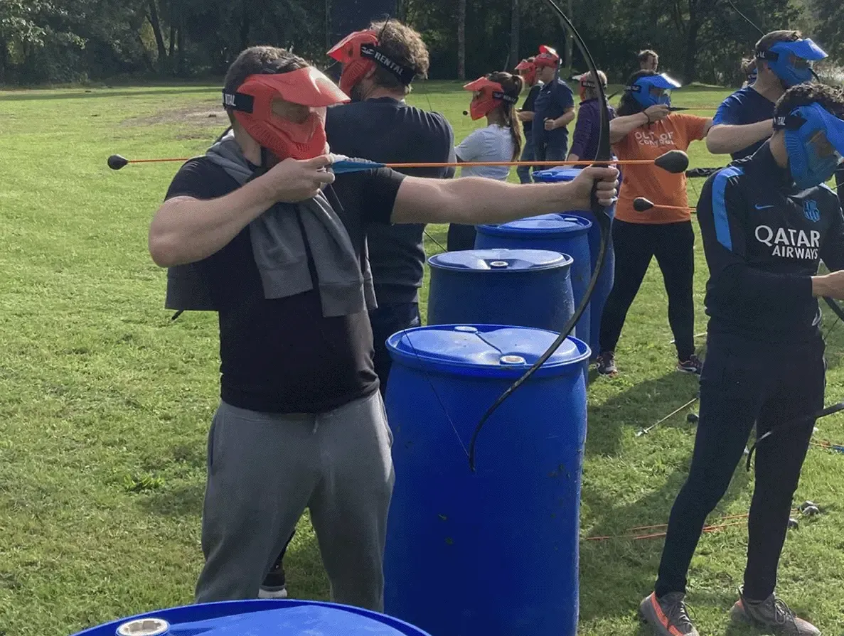 4everyware medewerkers nemen deel aan een teambuilding activiteit boogschieten, met rode en blauwe beschermende maskers en pijlen op een grasveld.