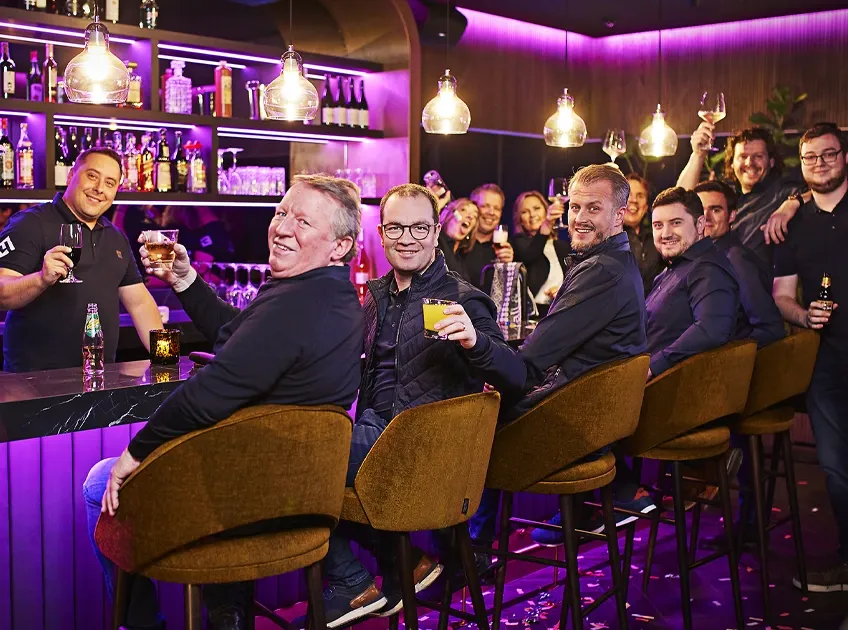 Teamfoto van medewerkers in zwarte kleding die samen proosten met een glas in de hand