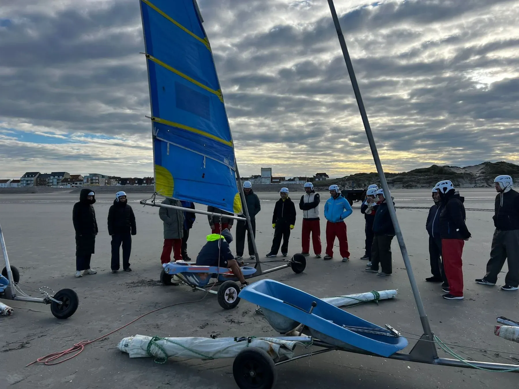 Groep mensen op het strand met veiligheidshelmen, aandachtig luisterend naar de uitleg van een instructeur voorafgaand aan een blowkarten activiteit